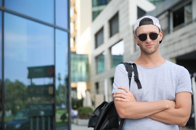 Photo of Handsome young man with stylish sunglasses and backpack near building outdoors, space for text