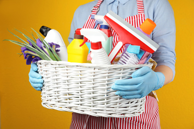 Photo of Woman holding basket with spring flowers and cleaning supplies on yellow background, closeup