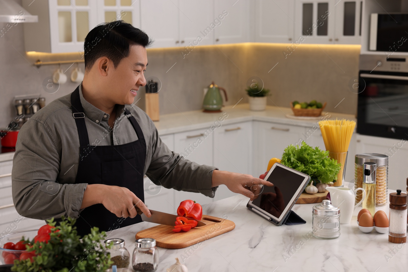 Photo of Cooking process. Man using tablet while cutting fresh bell pepper at countertop in kitchen