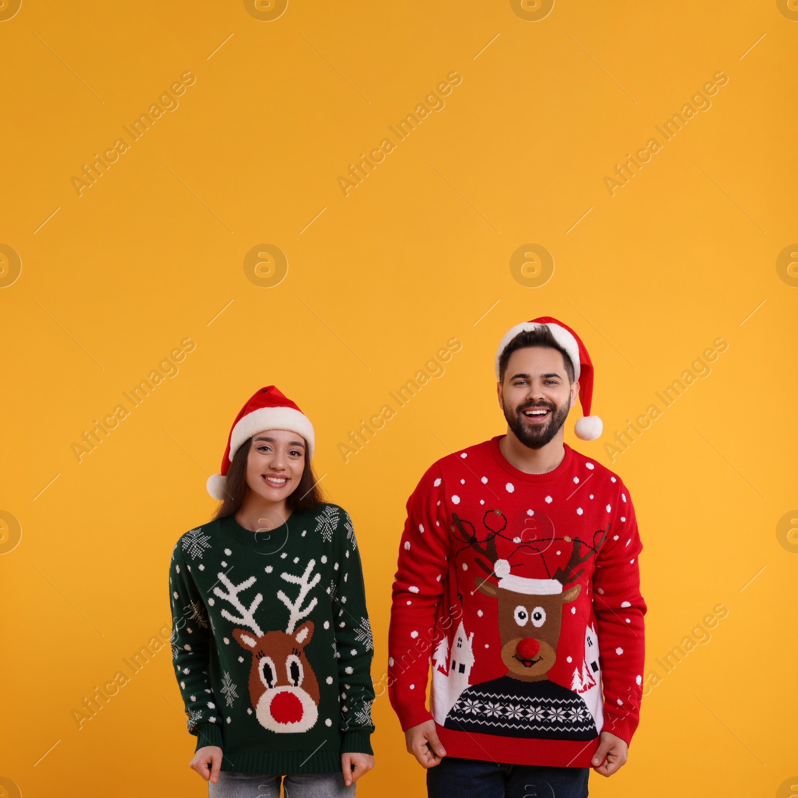 Photo of Happy young couple in Santa hats showing Christmas sweaters on orange background. Space for text