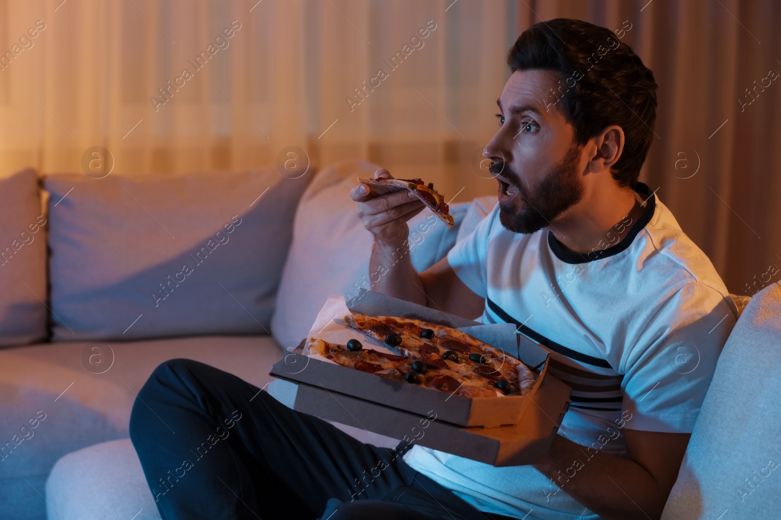 Photo of Man eating pizza while watching TV on sofa at night. Bad habit