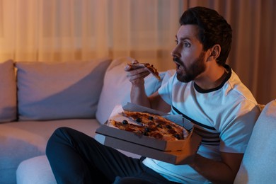 Photo of Man eating pizza while watching TV on sofa at night. Bad habit
