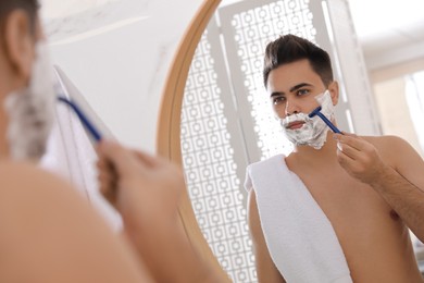 Photo of Handsome young man shaving with razor near mirror in bathroom