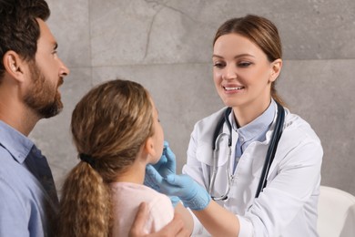 Photo of Smiling doctor examining girl`s oral cavity near her father indoors