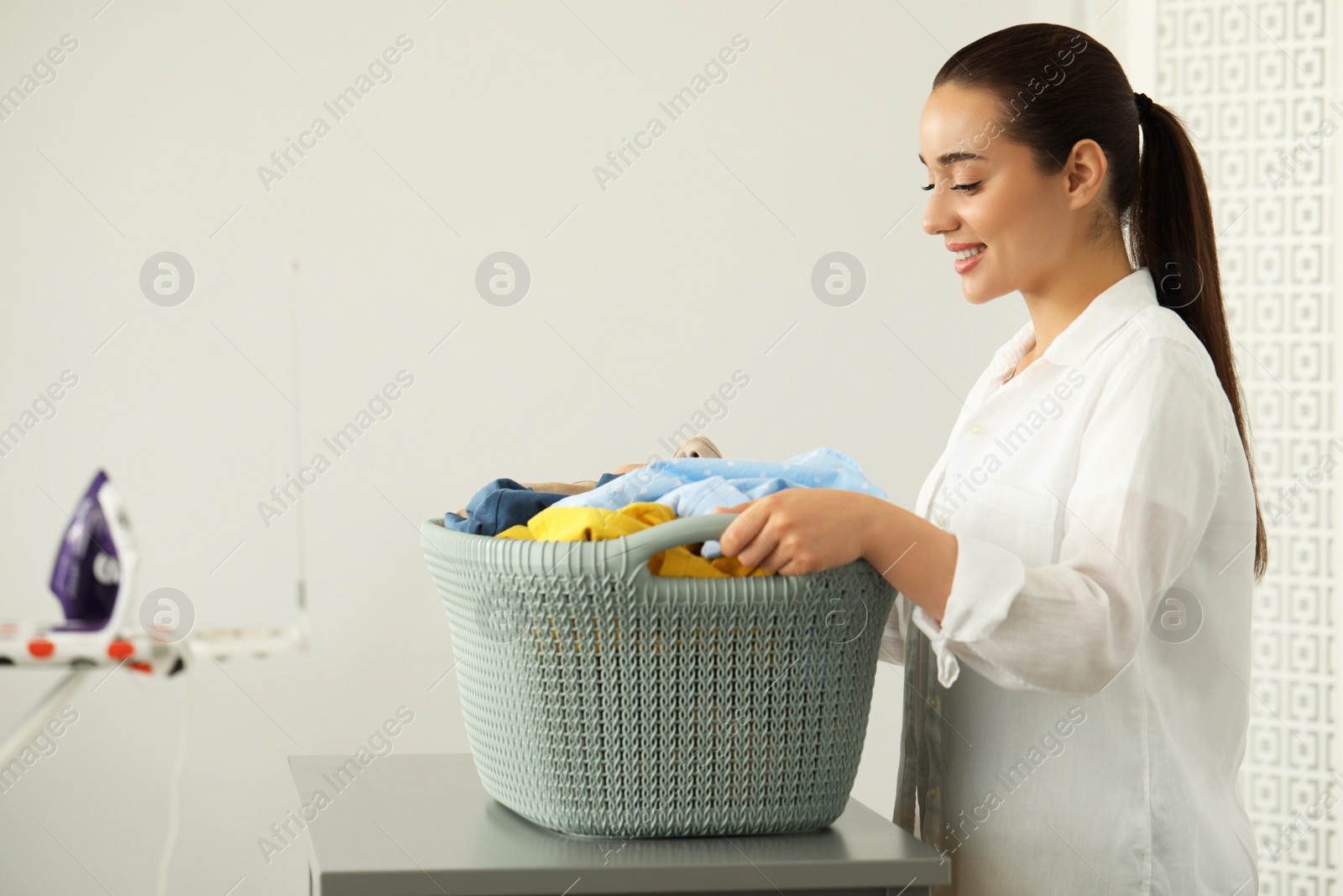 Photo of Young woman with basket full of clean laundry at table indoors