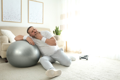 Photo of Lazy overweight man with sport equipment on floor at home