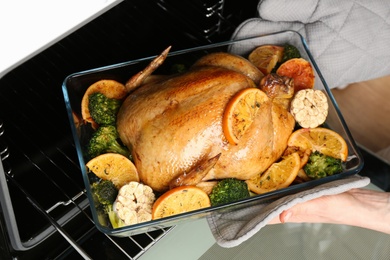 Woman taking baked chicken with oranges and vegetables out of oven, closeup