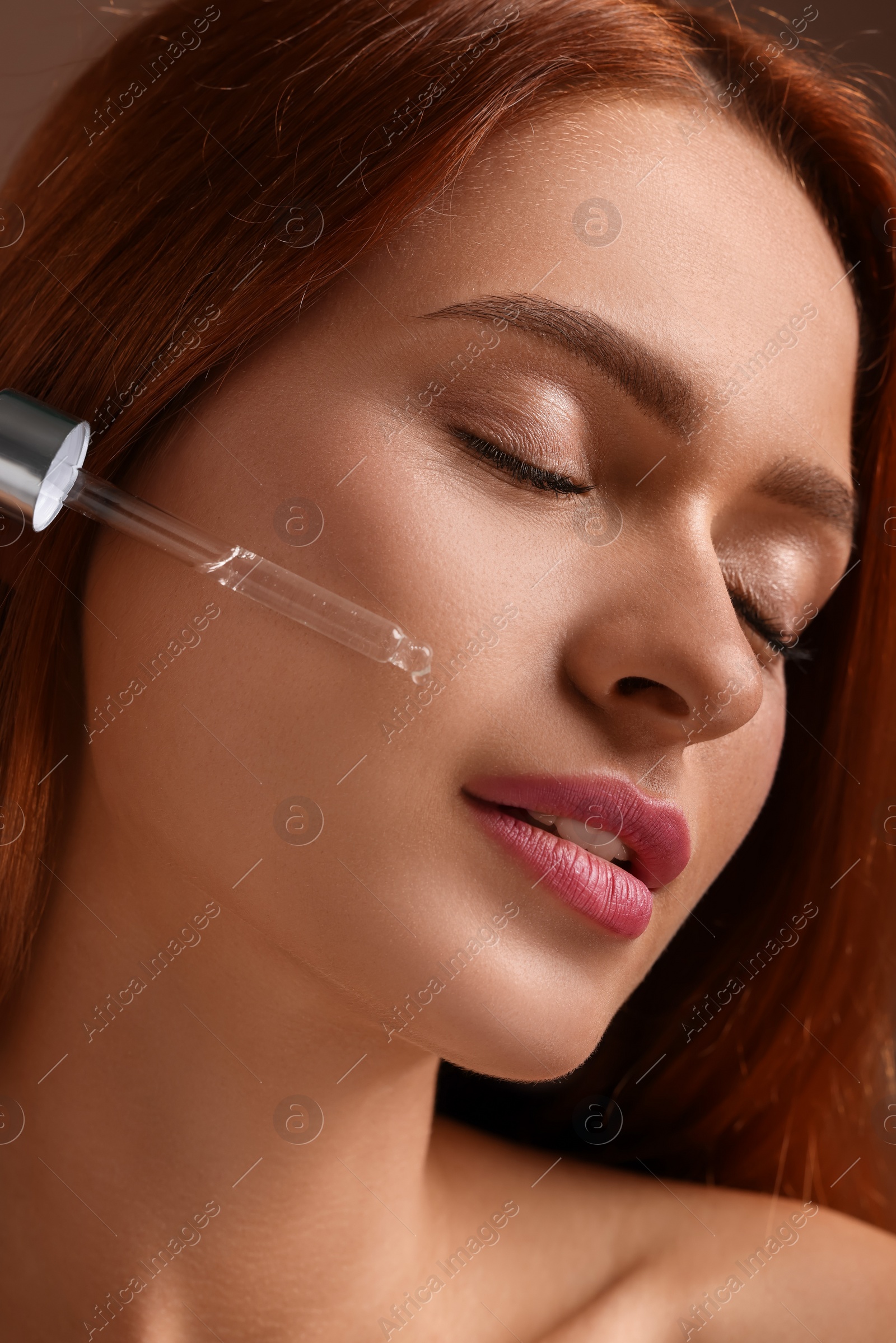 Photo of Beautiful young woman applying cosmetic serum onto her face on brown background, closeup