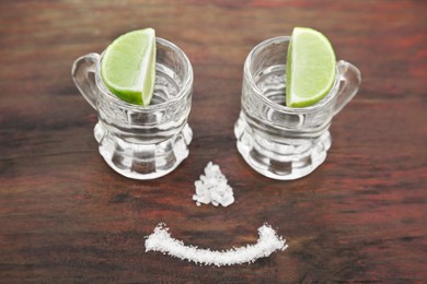 Photo of Mexican tequila shots with lime slices and salt in form of smile on wooden table. Drink made from agave