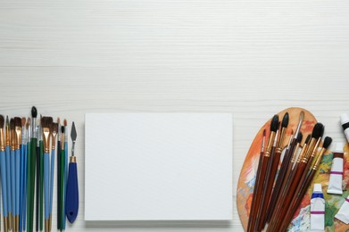 Blank canvas, tools, palette and paints on white wooden table, flat lay. Space for text