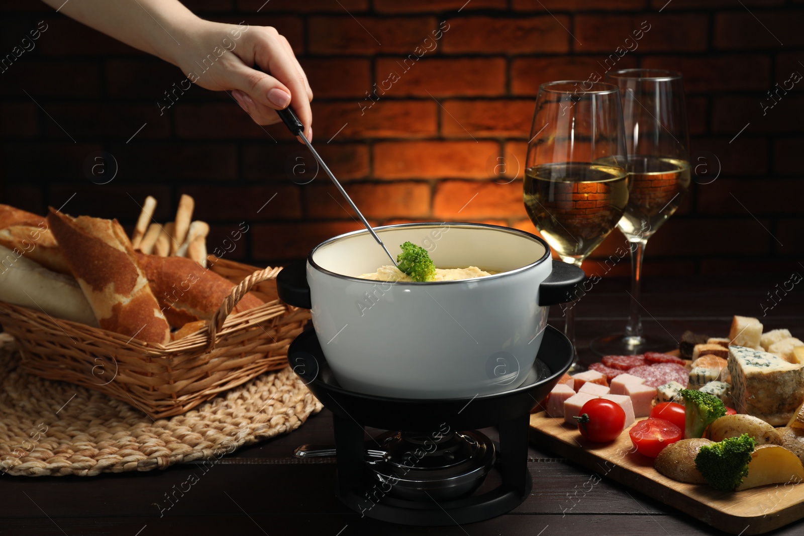 Photo of Woman dipping piece of broccoli into fondue pot with melted cheese at wooden table with wine and snacks, closeup