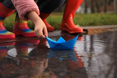 Photo of Little girl and her mother playing with paper boat near puddle outdoors, closeup