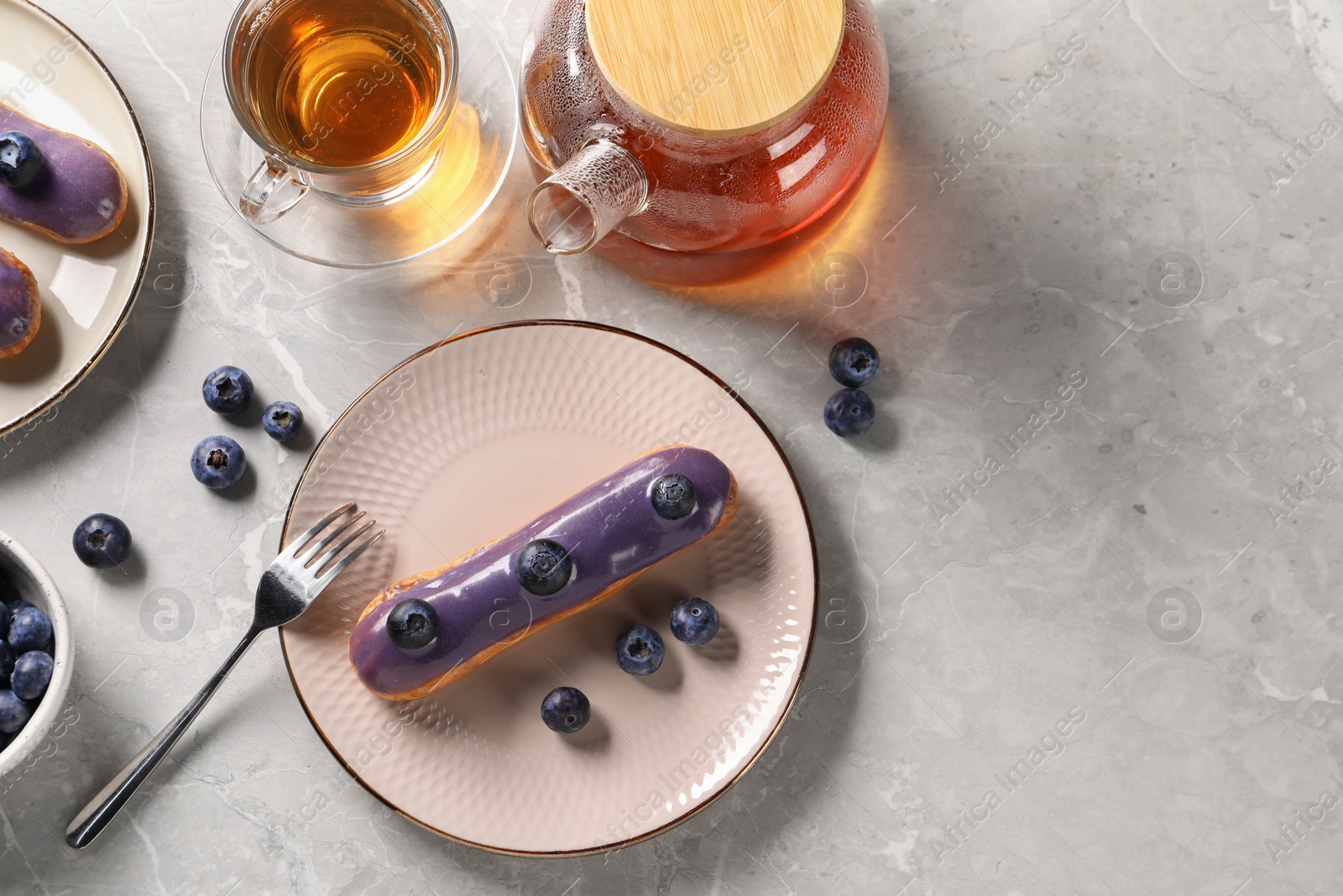 Photo of Tasty glazed eclair with blueberries and tea on grey marble table, flat lay. Space for text