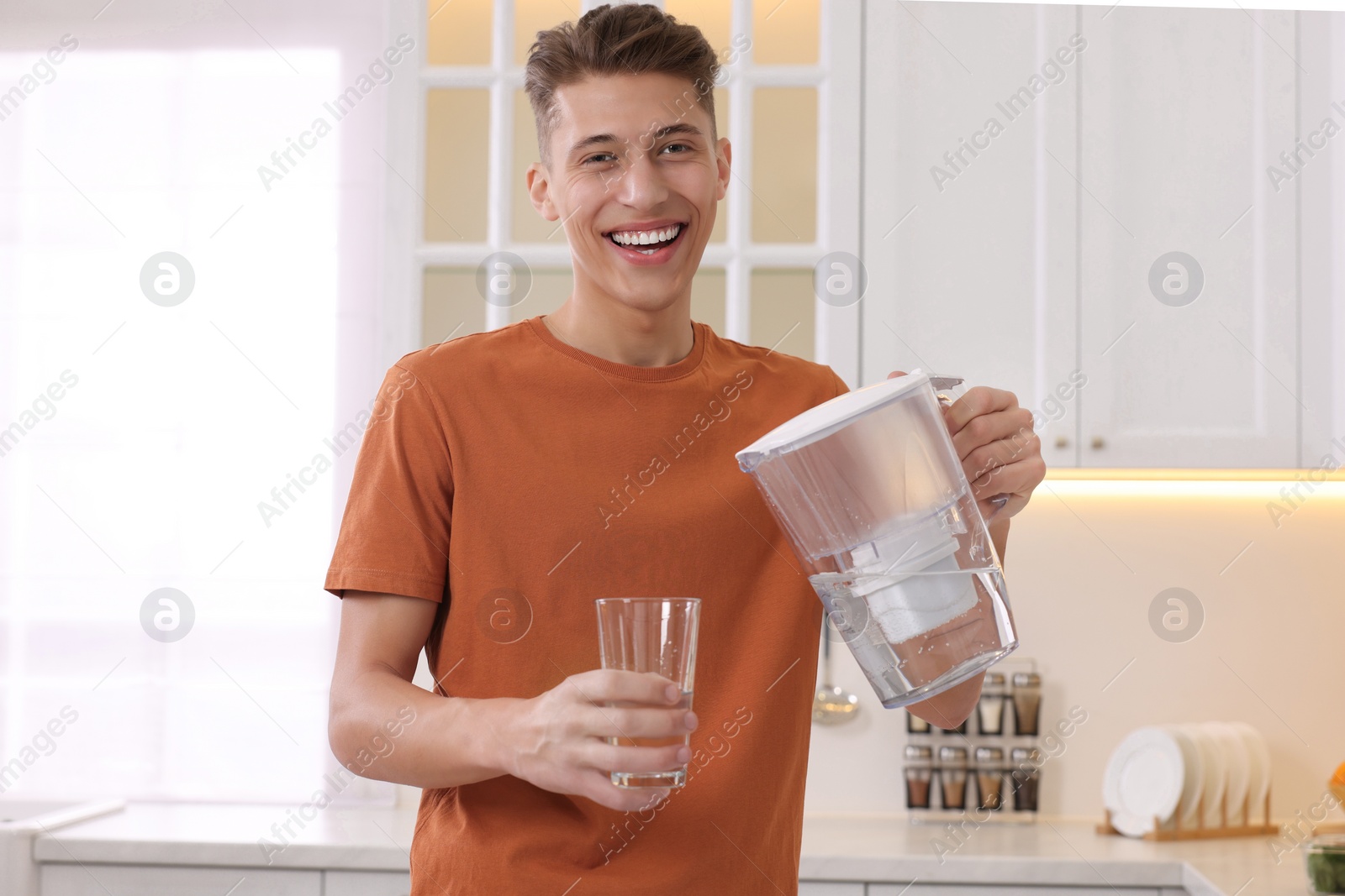 Photo of Happy man pouring water from filter jug into glass in kitchen