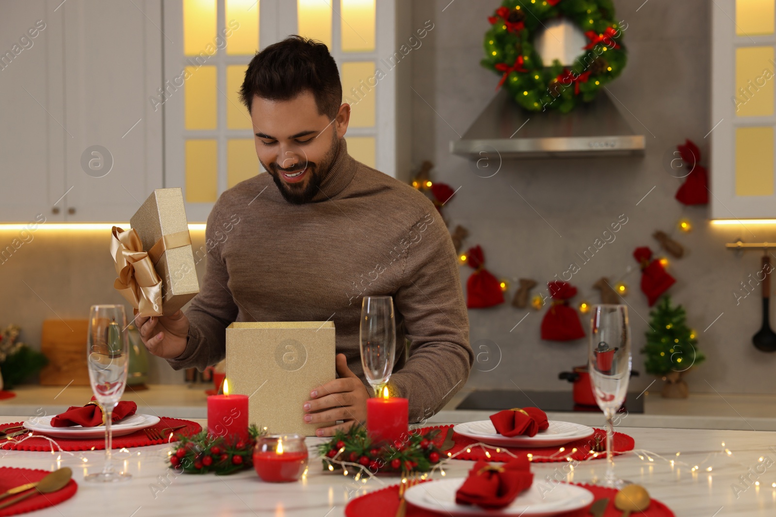 Photo of Happy young man opening Christmas gift at table in kitchen