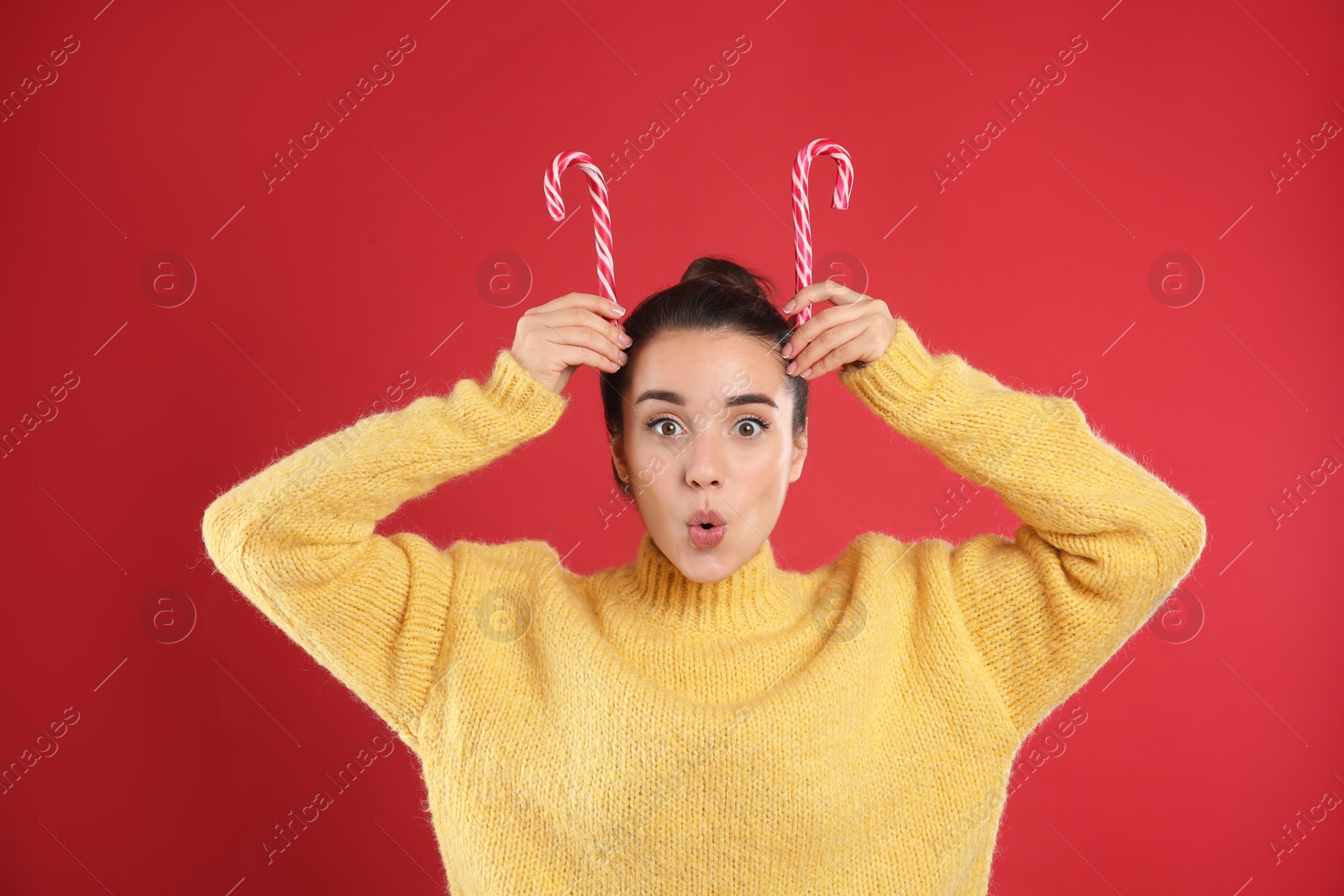 Photo of Surprised young woman in yellow sweater holding candy canes on red background. Celebrating Christmas