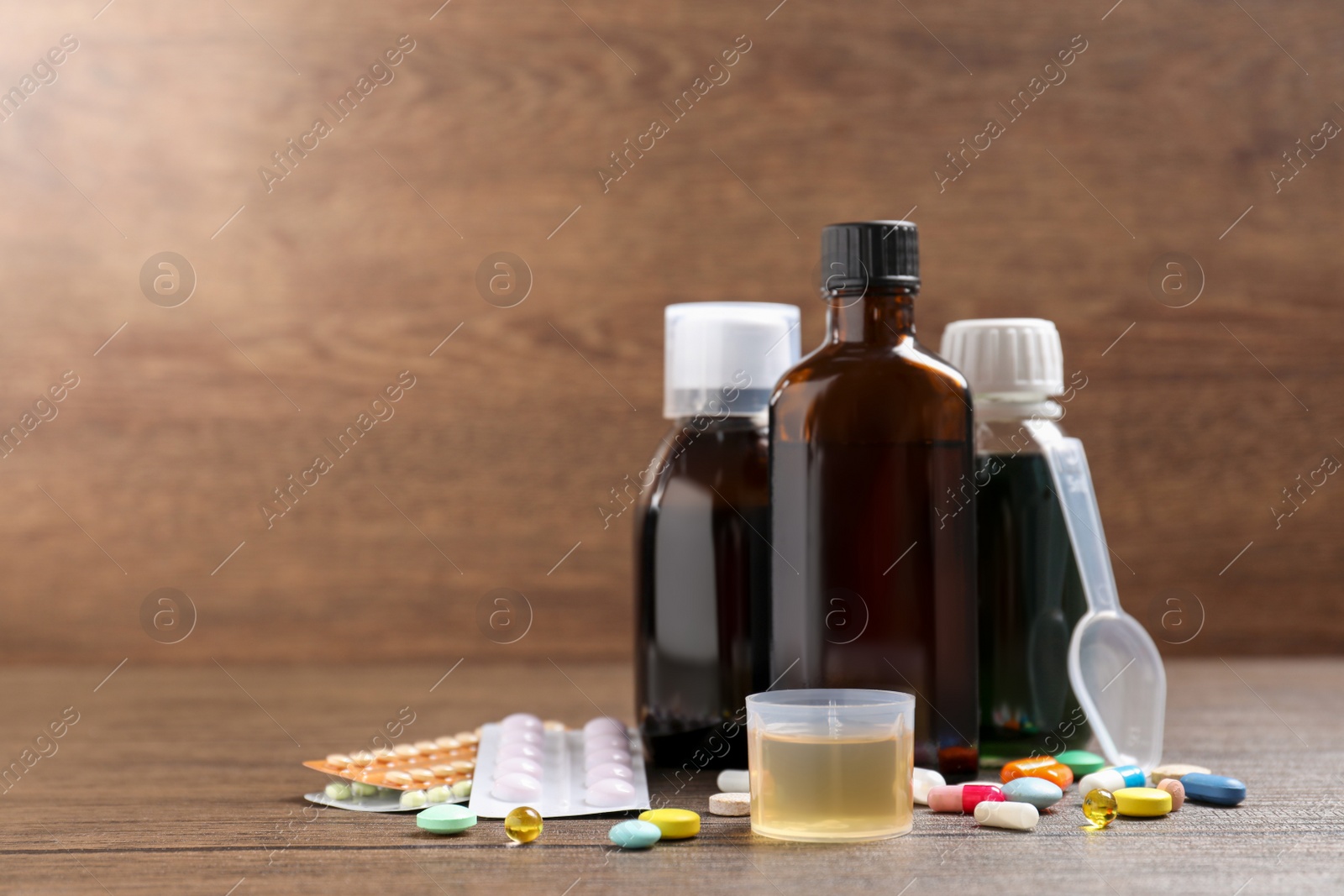 Photo of Bottles of syrup, measuring cup, dosing spoon and pills on wooden table, space for text. Cold medicine