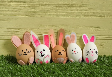 Photo of Easter eggs as cute bunnies on green grass against yellow wooden background