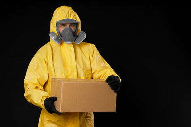 Photo of Man wearing chemical protective suit with cardboard box on black background, space for text. Prevention of virus spread