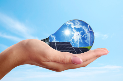 Image of Alternative energy source. Woman holding light bulb with solar panels and wind turbines, closeup
