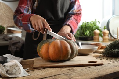 Photo of Woman cutting fresh ripe pumpkin at wooden table in kitchen, closeup