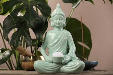 Buddhism religion. Decorative Buddha statue with burning candle on wooden table and monstera against light brown wall
