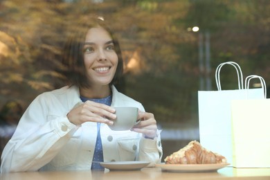 Photo of Special Promotion. Happy woman with shopping bags and cup of drink in cafe, view from outdoors