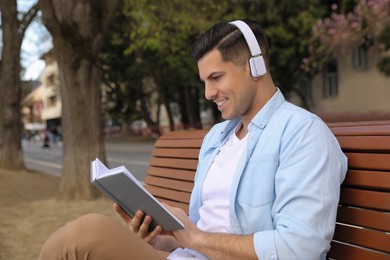 Photo of Happy man with headphones reading book on bench outdoors