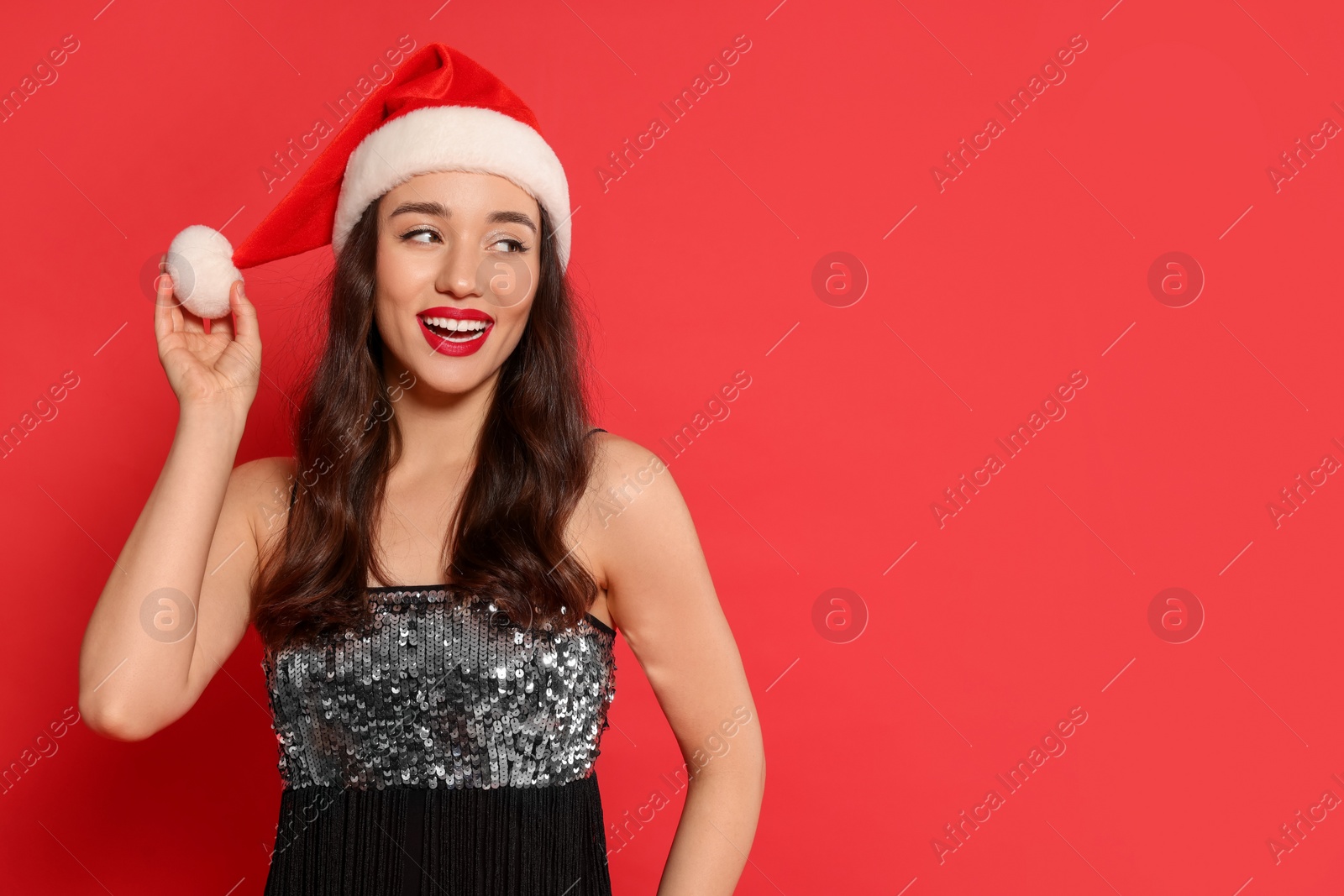 Photo of Christmas celebration. Beautiful young woman in stylish dress and Santa hat on red background, space for text