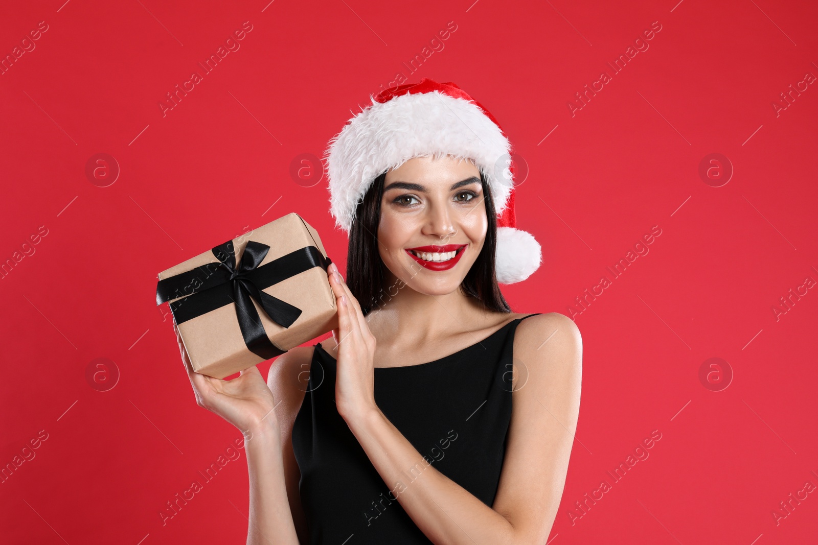 Photo of Woman in black dress and Santa hat holding Christmas gift on red background