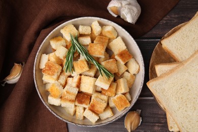 Delicious crispy croutons with rosemary in bowl, toast bread slices and garlic on wooden table, flat lay