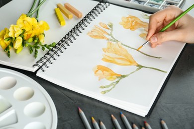 Photo of Woman drawing beautiful freesia flowers in sketchbook at black table, closeup