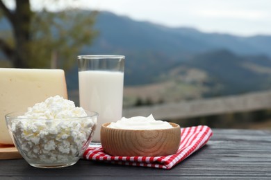 Photo of Tasty cottage cheese and other fresh dairy products on black wooden table in mountains. Space for text