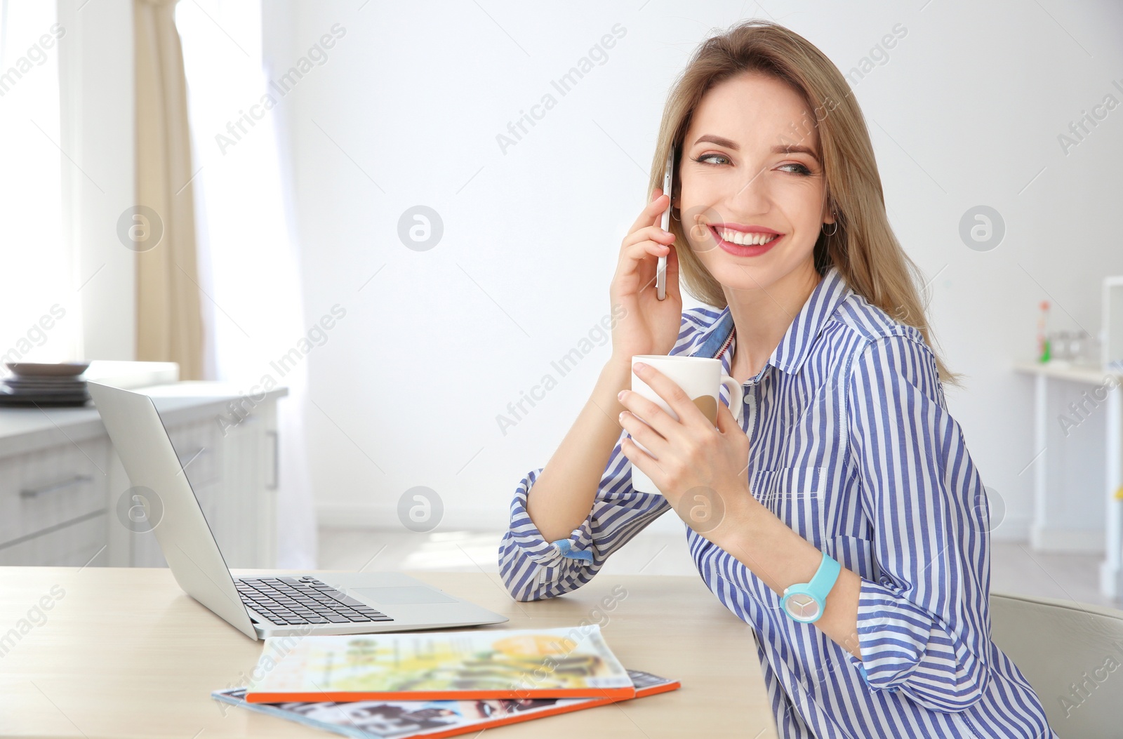 Photo of Young woman talking on phone while using laptop indoors