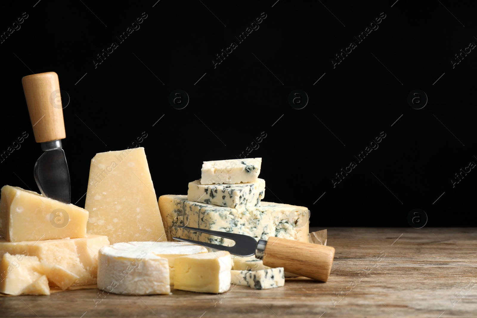 Photo of Different sorts of cheese, fork and knife on wooden table against black background. Space for text