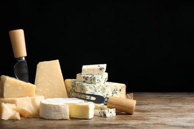 Different sorts of cheese, fork and knife on wooden table against black background. Space for text