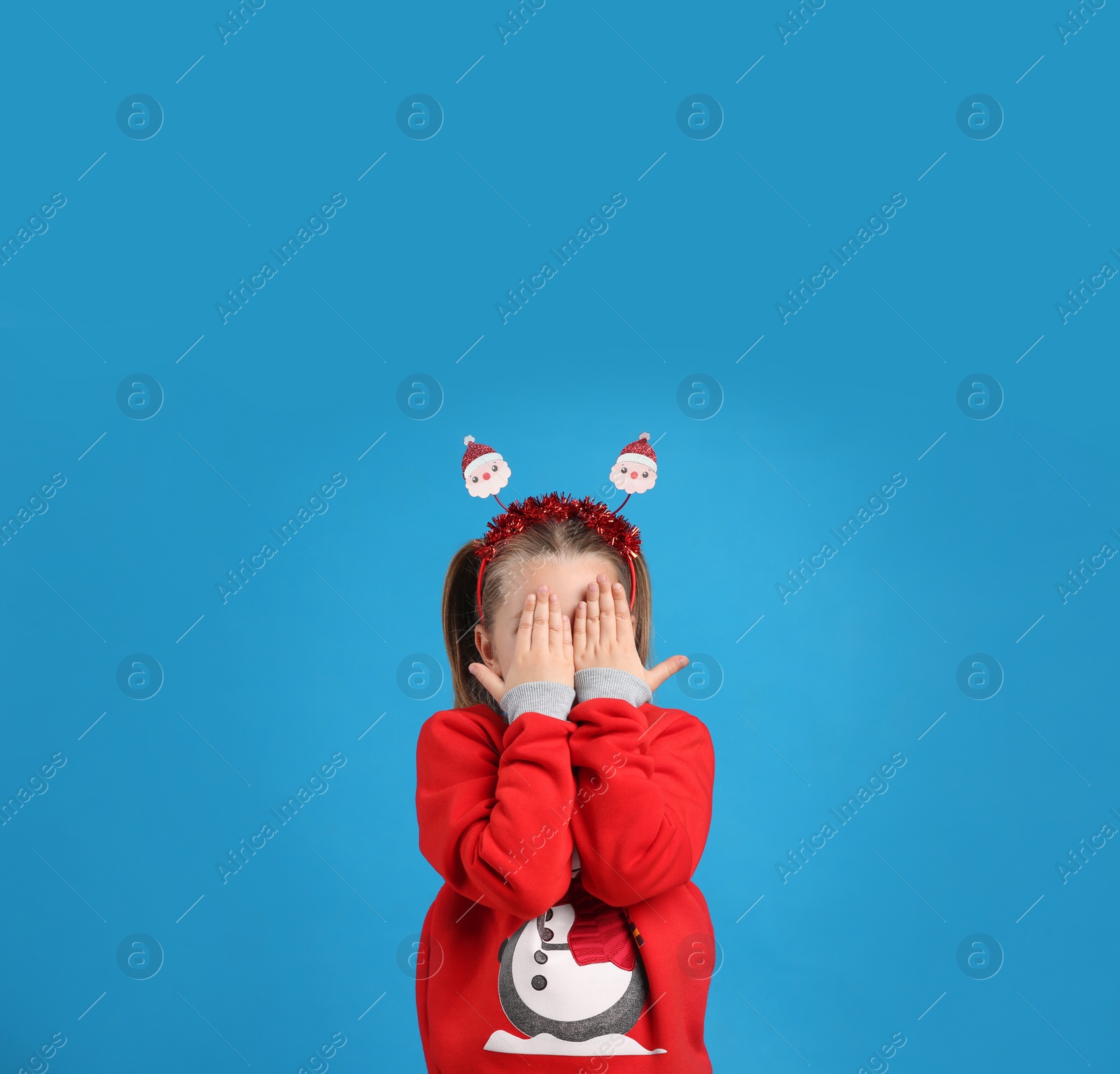 Photo of Cute little girl in Christmas sweater and Santa headband hiding her face on blue background