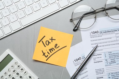 Photo of Reminder note with words Tax Time, documents, computer keyboard and calculator on grey table, flat lay