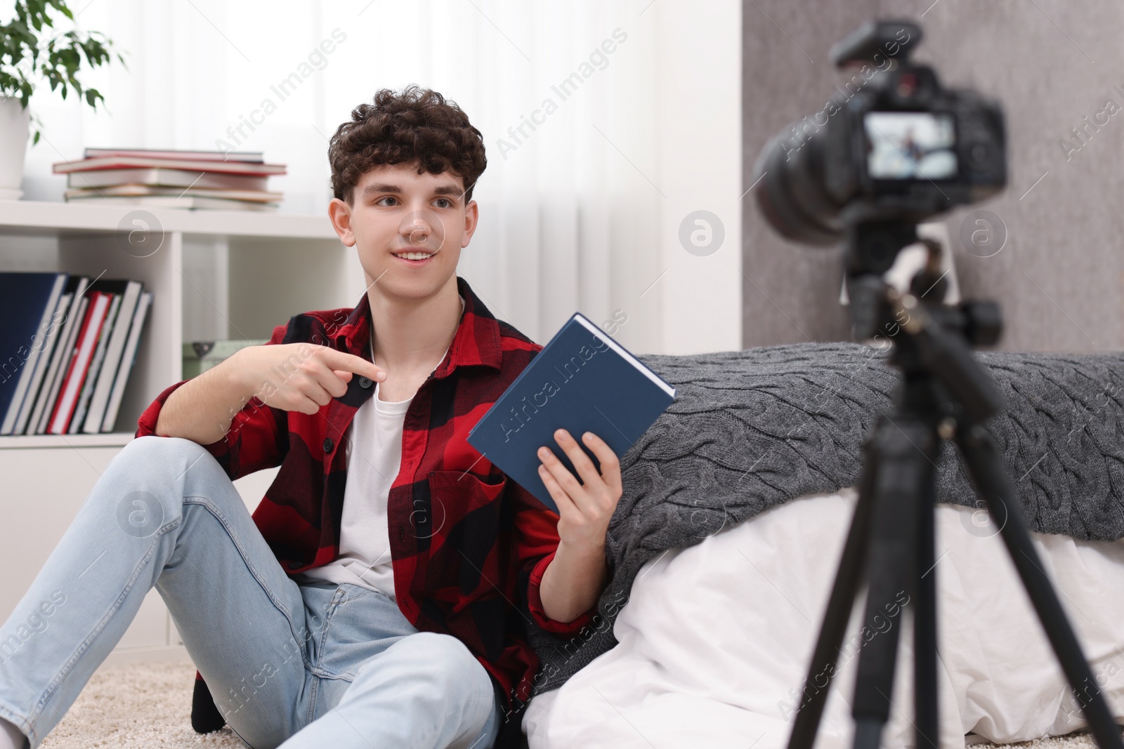 Photo of Smiling teenage blogger pointing at book while streaming at home