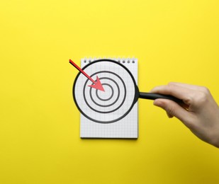 Image of Woman holding magnifying glass over notebook on yellow background, top view. Illustration of target and arrow