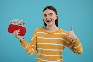 Photo of Happy woman holding wallet with dollar banknotes and showing thumbs up on light blue background