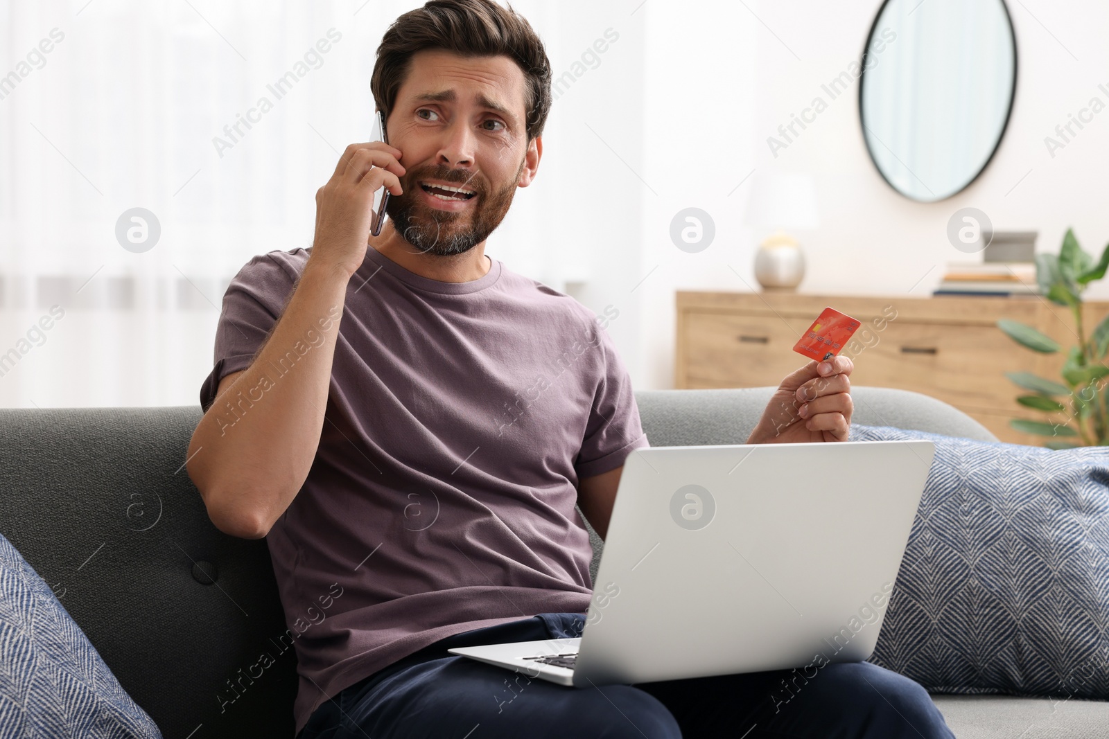 Photo of Emotional man with credit card and laptop talking on phone at home. Be careful - fraud