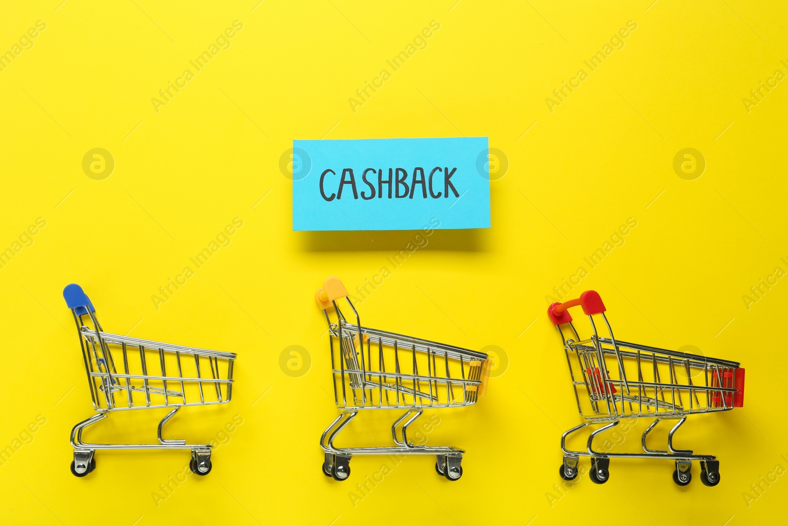 Photo of Card with word Cashback and shopping carts on yellow background, flat lay