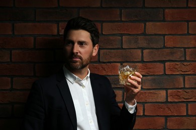 Photo of Handsome man in formal suit holding glass of whiskey near red brick wall. Space for text