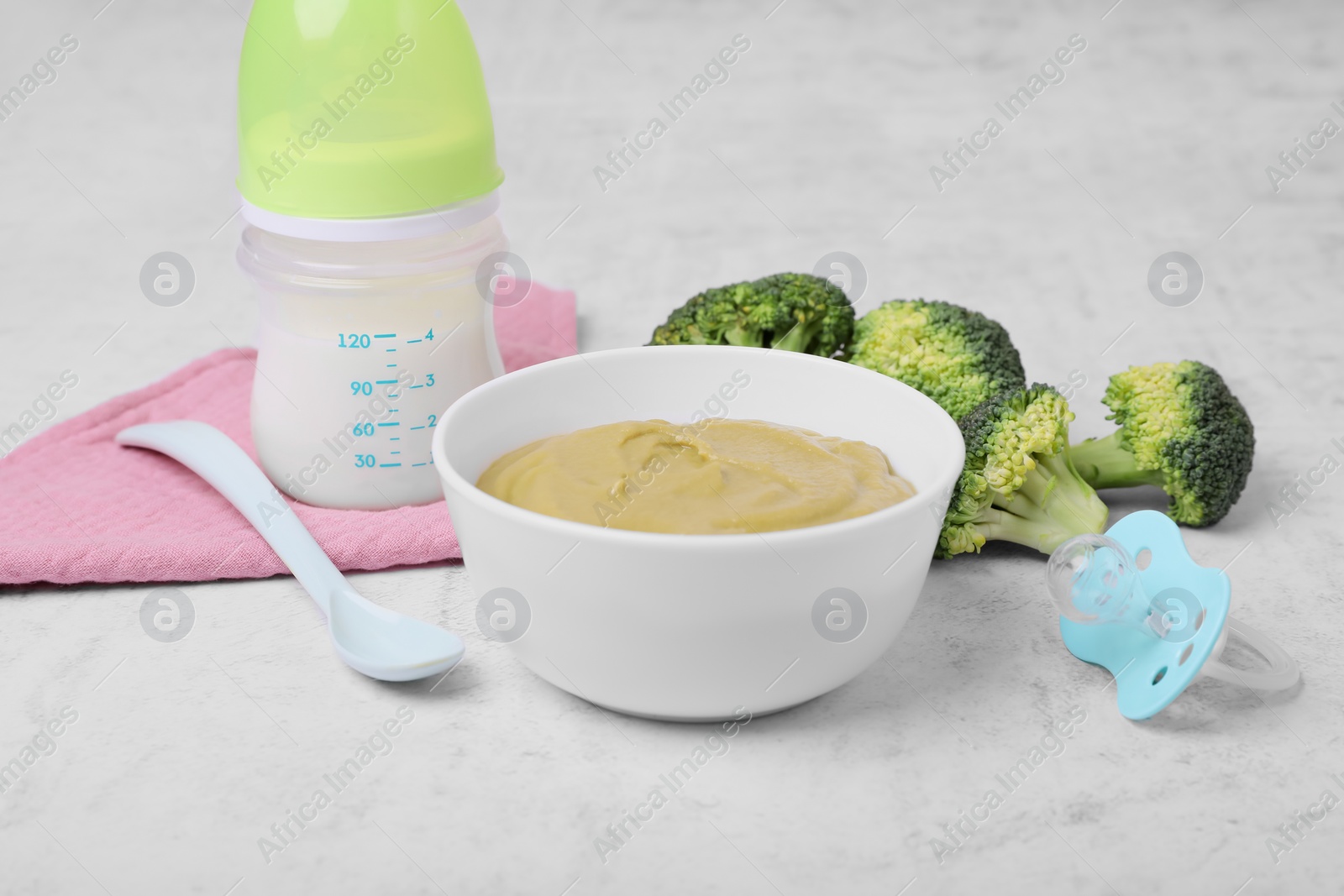 Photo of Bowl with healthy baby food, broccoli, spoon, pacifier and bottle of milk on white table