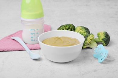 Photo of Bowl with healthy baby food, broccoli, spoon, pacifier and bottle of milk on white table