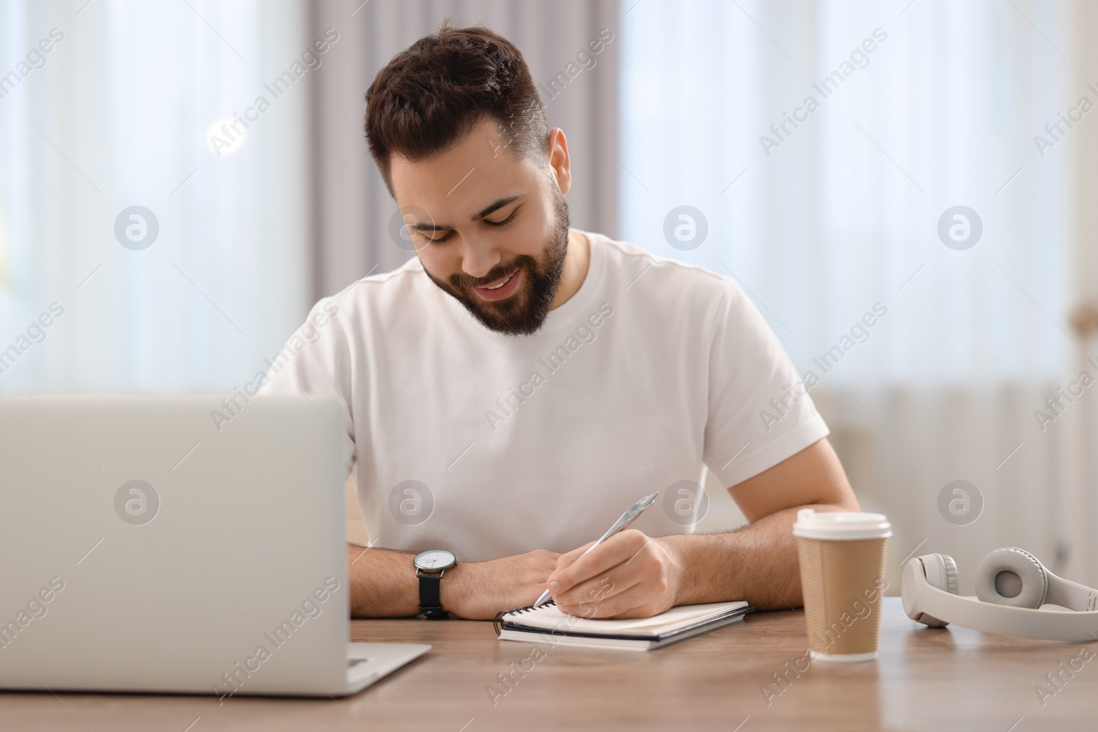 Photo of Young man writing down notes during webinar at table in room