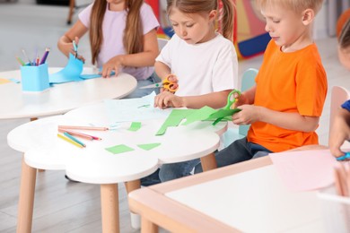 Photo of Cute little children cutting color paper with scissors at desks in kindergarten. Playtime activities