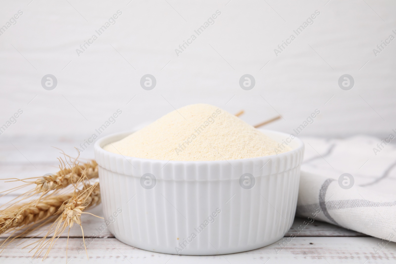 Photo of Uncooked organic semolina in bowl and spikelets on white wooden table, closeup