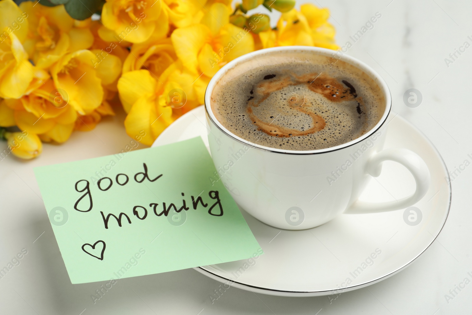 Photo of Cup of aromatic coffee, beautiful yellow freesias and Good Morning note on white marble table